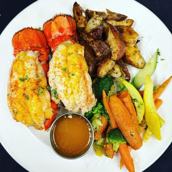 Two lobster tails with roasted potatoes, mixed vegetables, and a side of butter on a white plate.