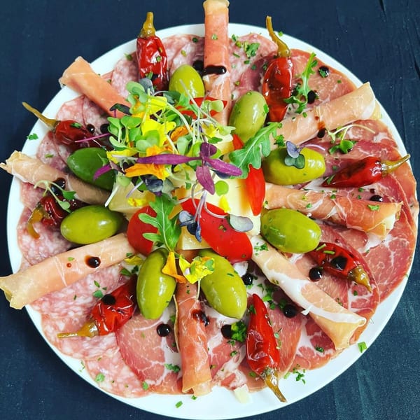 A charcuterie board with various meats, olives, peppers, and a colorful microgreen salad.