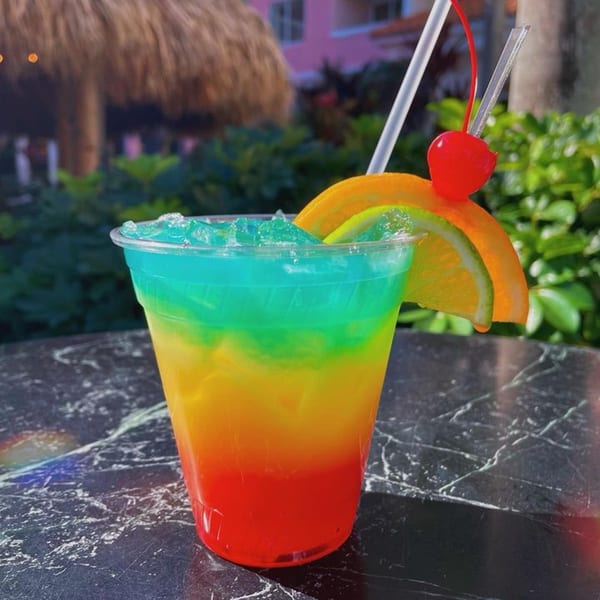A vibrant, layered tropical drink with blue, green, yellow, and red, garnished with fruit and a cherry.