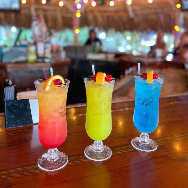 Three colorful cocktails, red, yellow, and blue, garnished with fruit on a bar at The Islander Grill.