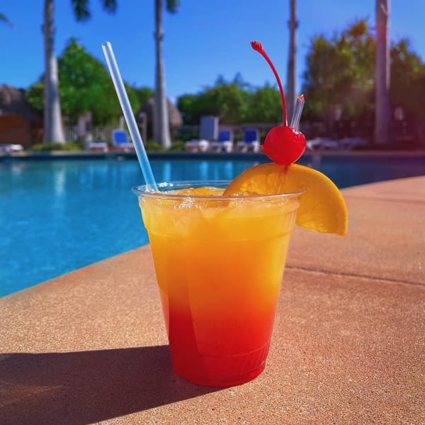 Tropical drink with orange and red layers, garnished with an orange slice and cherry, by a pool.